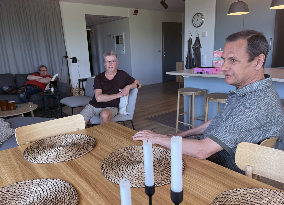 En man halvligger i en soffa längst t v i bilden. I motten sitter en man i en fåtölj. Till höger sitter en man vid ett köksbord, med vänster hand på bordet.