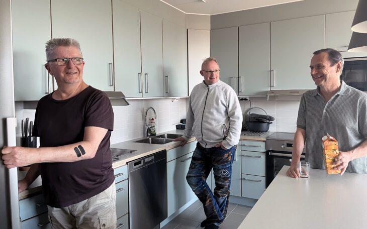 Tre män i ett kök. Längst t v i bilden syns en man i grått hår och glasögon som är i färd med att öppna en kylskåpsdörr. I mitten, i kökets hörn, står en kortklippt man med glasögon. Han håller ena handen i fickan och den andra på köksbänken. Längst tv en kortklippt man med mörkt hår som står vid köksön. Han ska precis hälla upp ett glas apelsinjuice. Alla tre ser glada ut.