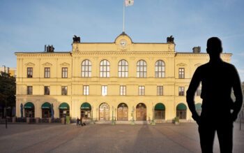 Bild på en stor, gul byggnad i sten. Det är ett vackert solljus i bilden. I bilden finns även silhuetten av en man inflikad i ena hörnet. Silhuetten går helt i svart.