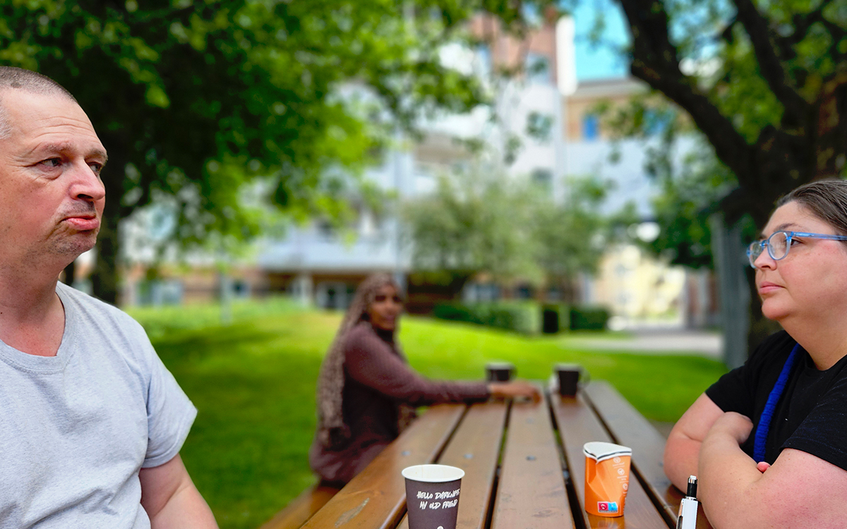 En man och en kvinna i trädgårdsmiljö. De sitter på varsin sida om ett bord. I bakgrunden syns en annan kvinna, men den delen av bilden är medvetet oskarp.