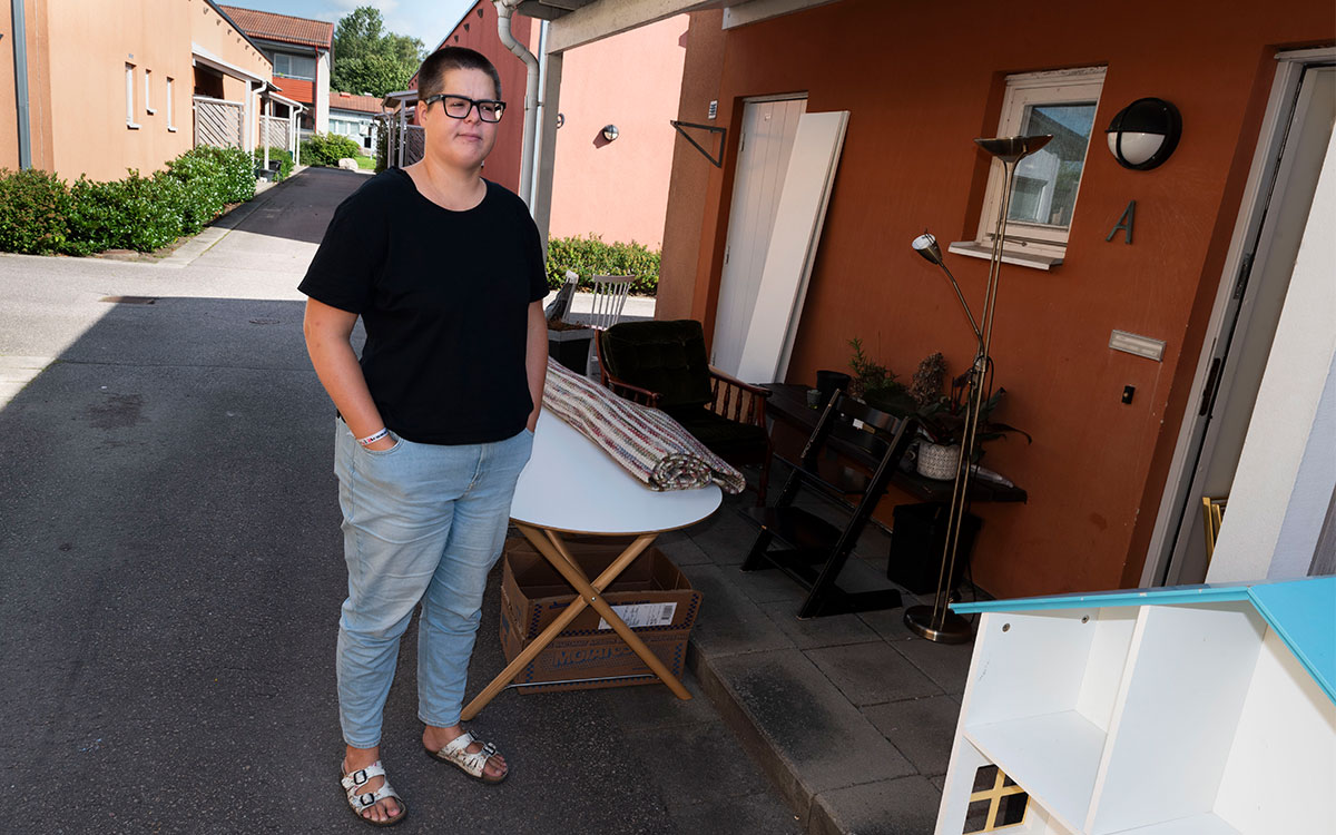 En ung kvinna i kort hår och glasögon som står framför ett hus där det står diverse möbler och annat bohag.