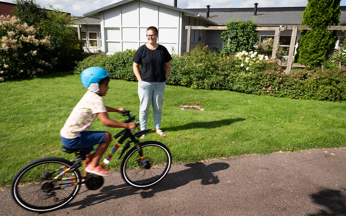 En ung kvinna i kort hår och glasögon som tittar på när en pojke cyklar förbi på en cykelväg framför en gräsmatta.
