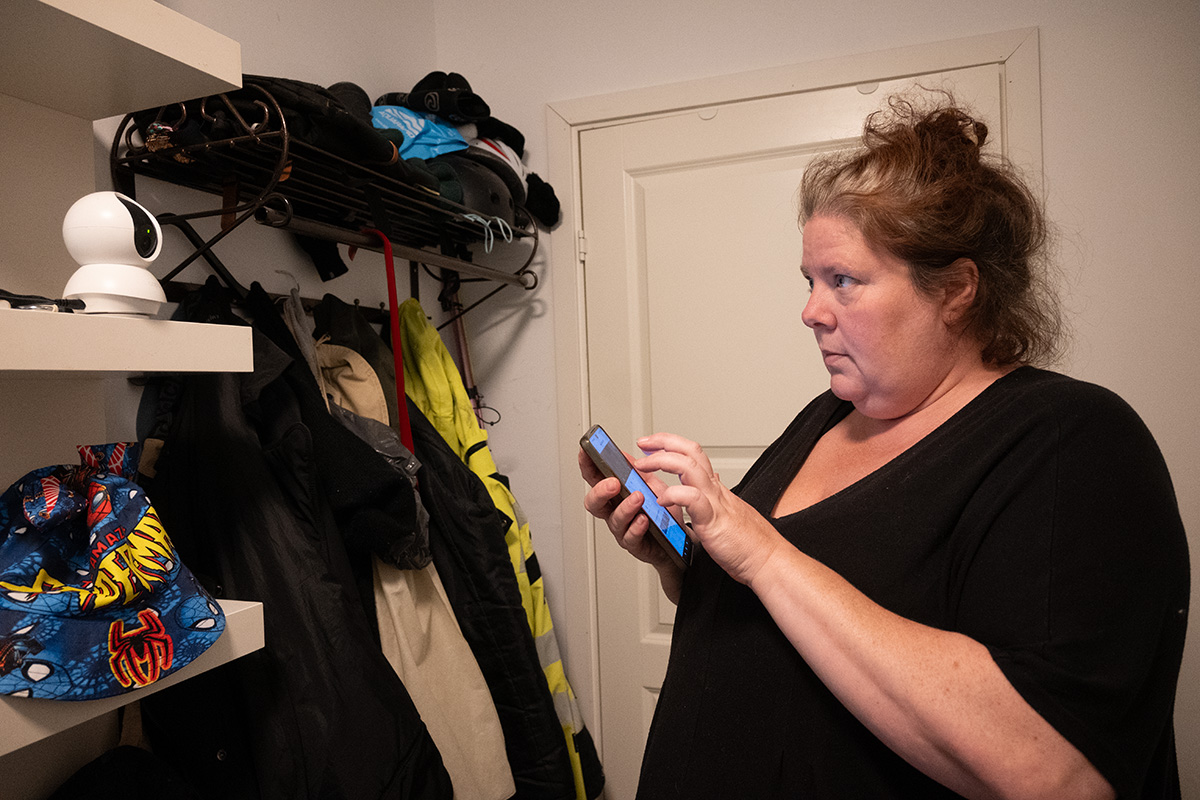 En kvinna med en mobil i handen. Tittar på en övervakningskamera som står på en hylla.