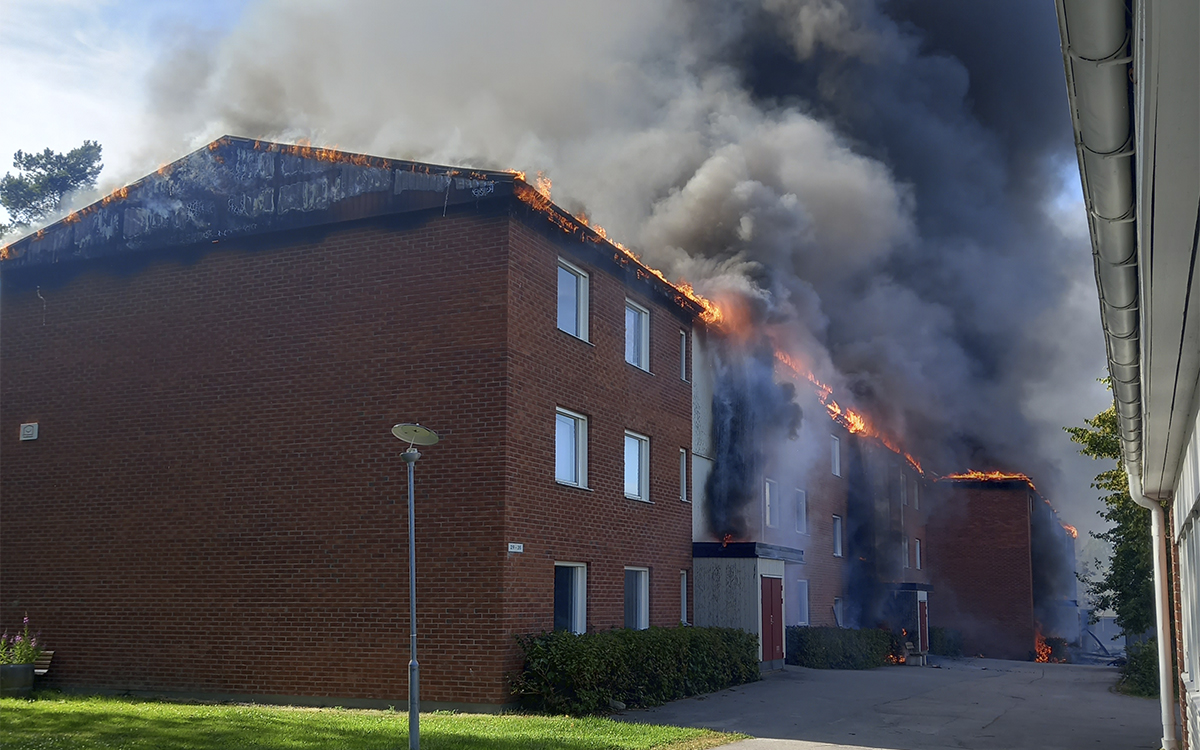 Eldslågor slår upp från taket på ett trevåningshus i tegel och luften ovanför huset är fylld av grå och svart brandrök. Det ser också ut att vara eldslågor och svart brandrök längs med trappuppgångarna. I förgrunden en grön gräsmatta. Branden på Stormyravägen i Hudiksvall spred sig mycket snabbt upp på vinden och sen ner i alla fyra trappuppgångar. 36 lägenheter förstördes i branden.