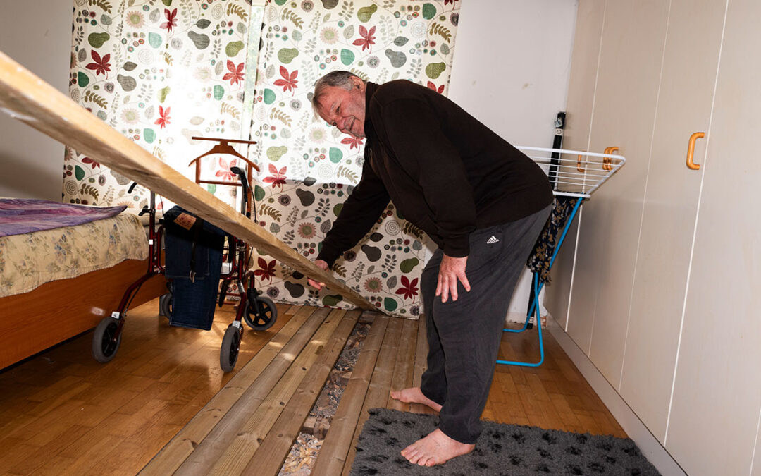 En äldre man som står i ett rum bredvid en rullator och håller i en lös planka från en övertäckning av ett hål i parkettgolvet.