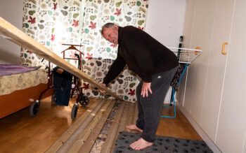 En äldre man som står i ett rum bredvid en rullator och håller i en lös planka från en övertäckning av ett hål i parkettgolvet.