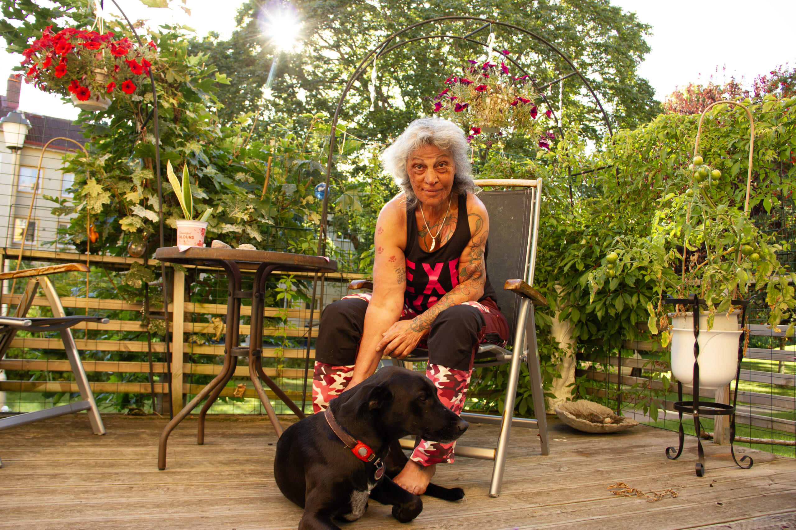 Zahra Medzeta i svart linne med rosaröda stora bokstäver ("EX") och byxor med rödrosakamouflagemönster sitter på en solstol på altanen, med grönska i bakgrunden. Framför henne ligger hunden ZeUs, som har mörk päls, och tittar åt sidan. Zahra Medzeta har fått veta att Uppsalahem inte kommer godkänna andrahandsuthyrning igen, vilket försvårar hennes vintrar utomlands.
