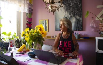 Zahra Medzeta sitter vid kökbordet med sin laptop framför sig och tittar ut genom fönstret till vänster i bild. På bordet en rosa- och vitrutig duk och en vas med solrosor, bakom henne en rosa vägg och en stor tavla med en kvinna i gråskala.