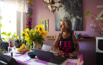 Zahra Medzeta sitter vid kökbordet med sin laptop framför sig och tittar ut genom fönstret till vänster i bild. På bordet en rosa- och vitrutig duk och en vas med solrosor, bakom henne en rosa vägg och en stor tavla med en kvinna i gråskala.