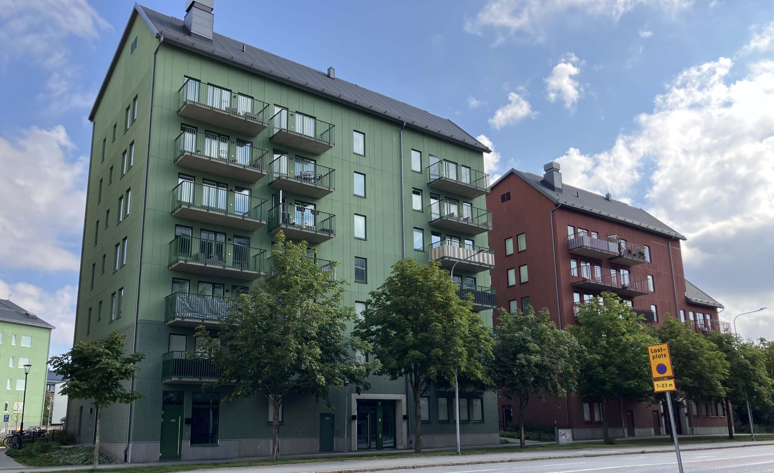 Två flerfamiljshus bredvid varandra. Det ena är grönt, det andra rött. Entréerna vetter mot en bilväg. Framför husen står gröna träd. 