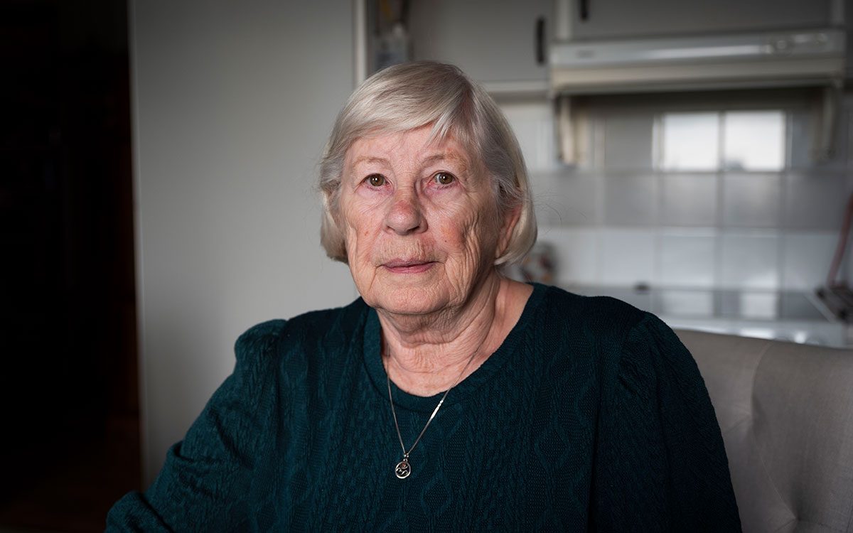 Ett porträtt på en äldre kvinna med bruna ögon och vitt hår som sitter i ett kök.