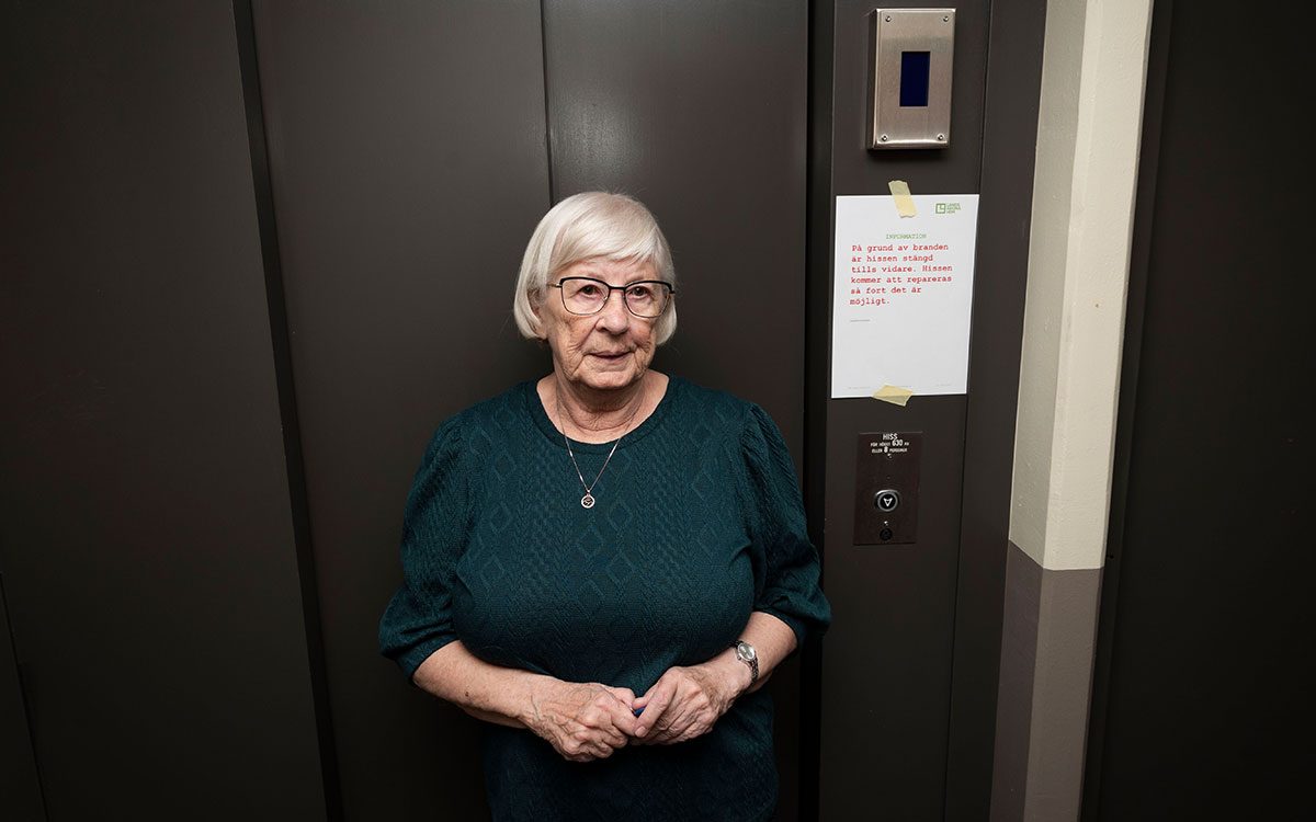 En äldre kvinna som står vid en hissdörr som har en lapp från Landskronahem där det står "På grund av branden är hissen avstängd tillsvidare".