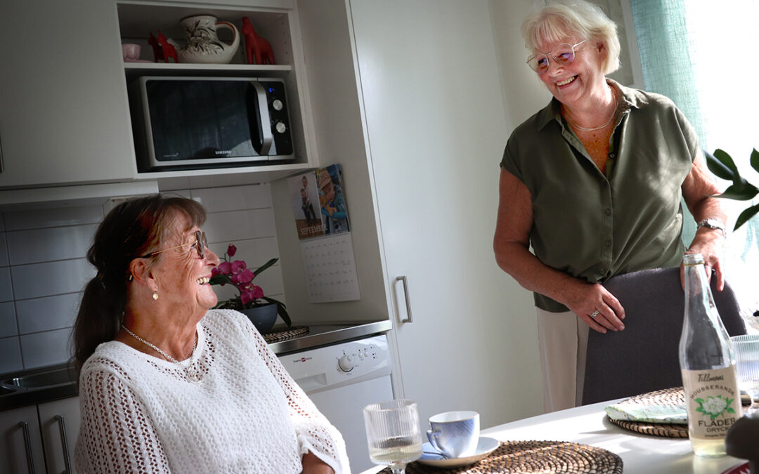 En kvinna sitter vid ett köksbord, en annan kvinna står upp. De tittar mot varandra och skrattar.