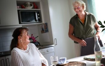 En kvinna sitter vid ett köksbord, en annan kvinna står upp. De tittar mot varandra och skrattar.