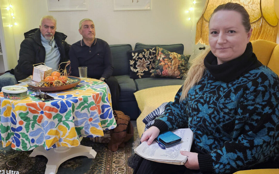 Tre personer i ett vardagsrum. I förgrunden i en fåtölj till höger sitter en kvinna med långt blont hår i en hästsvans på ryggen. Hon tittar allvarligt in i kameran. I händerna håller hon ett block med namnunderskrifter på, samt en mobil med turkos fodral. Hon har en stickad tröja med mönster i svart och turkos och en svart krage. Bakom henne syns en gul lampskärm. I soffan längs med väggen i bakgrunden sitter två män i medelåldern. Den ene har skägg och tittar in i kameran. Den andre tittar ut ur bilden. Framför dem ett bord med färgglad duk och en fruktskål på.