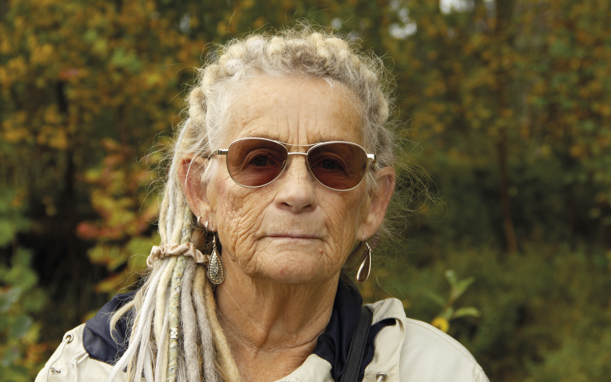 Porträtt på Aino Sundman i solglasögon ljus jacka och grå/vita/ljust färgade dreadlocks i en hästsvans. i bakgrunden träd i höstfärger.