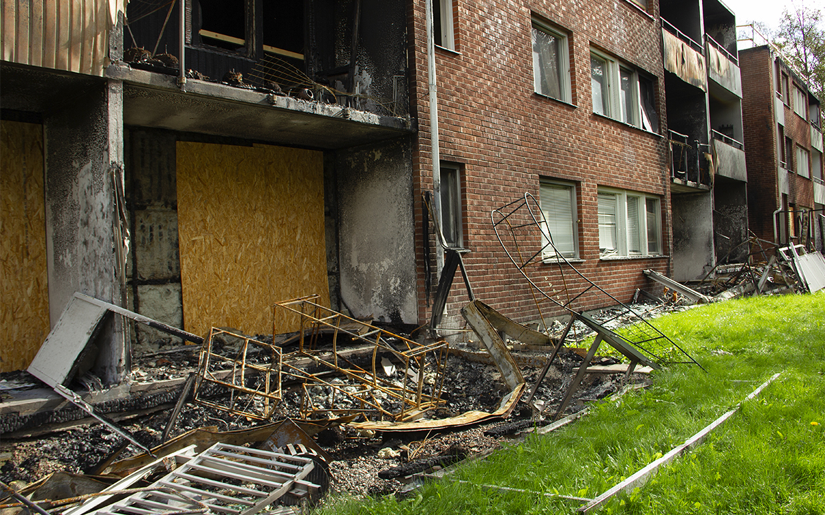 De två nedersta våningarna av ett svartbränt tegelhus, och intill det en grön gräsmatta. En svartbränd sned rosbåge halvligger bland annatbränt metallbråte på marken. Framför fönstret och dörren till uteplatsen närmast i bild sitter en masonitskiva. Branden på Stormyravägen i Hudiksvall spred sig via vinden. Ingen vet hur vanligt det är med bostadshus som saknar ordentliga brandceller på vinden.