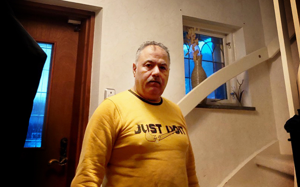 Man i gul tröja som tittar in i kameran. Han står bredvid en trappa men bara trappräcket syns.