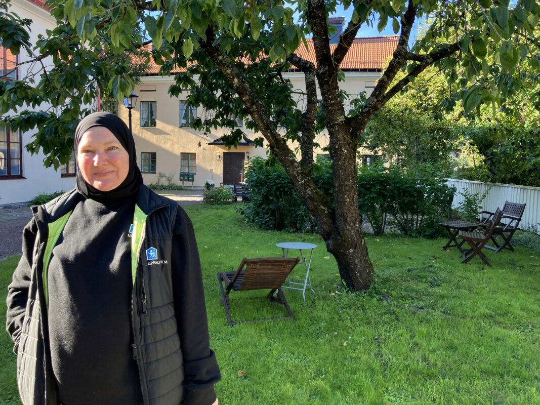 Kvinna med svart sjal och väst som det står Uppsalahem på. Hon står till vänster i bild. I bakgrunden en trädgård med ett träd, berså och trädgårdsstolar. Längst bort ett gulrappat hus.