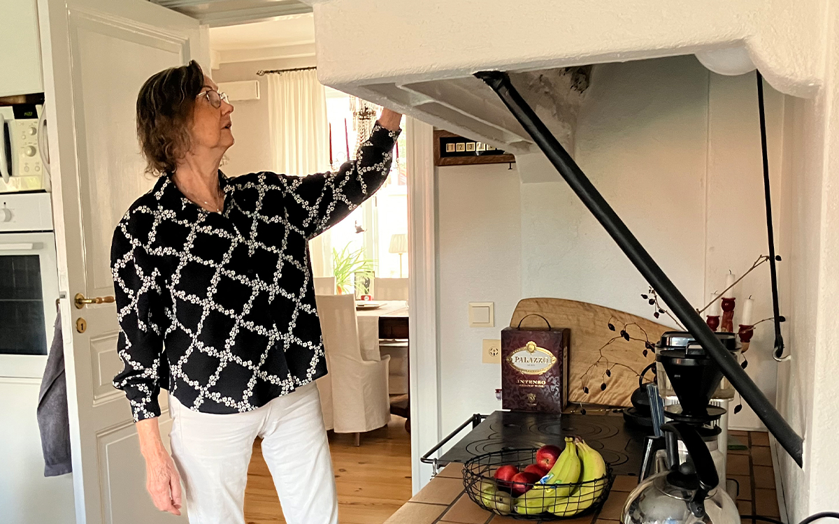 Kvinna 60+ framför en vedspis. Hon har svartvit mönstrad blus och vita byxor.