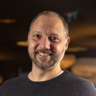 Porträttbild av en man som ler in i kameran. Han är tunnhårig och har ett mörkgrått kort skägg och mustasch. På sig har han en mörk t-shirt. Bakgrunden är mörk och suddig.