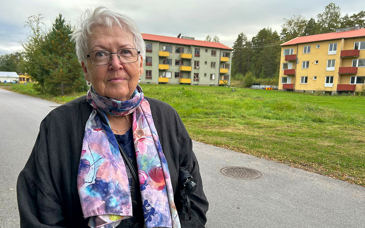 Kvinna som står vid en väg och tittar in i kameran. Bakom henne syns en gräsmatta och två flerfamiljshus.