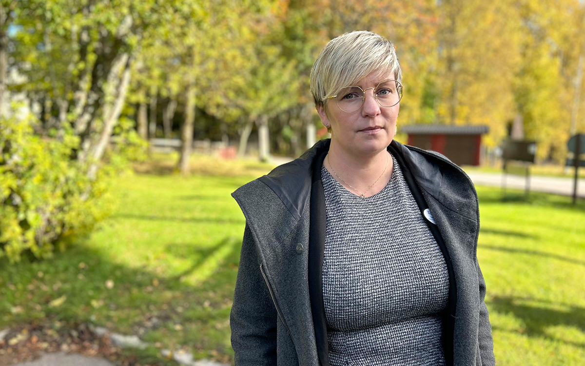 Kvinna med glasögon som tittar in i kameran. Bilden är tagen utomhus. En gräsmatta och träd syns i bakgrunden