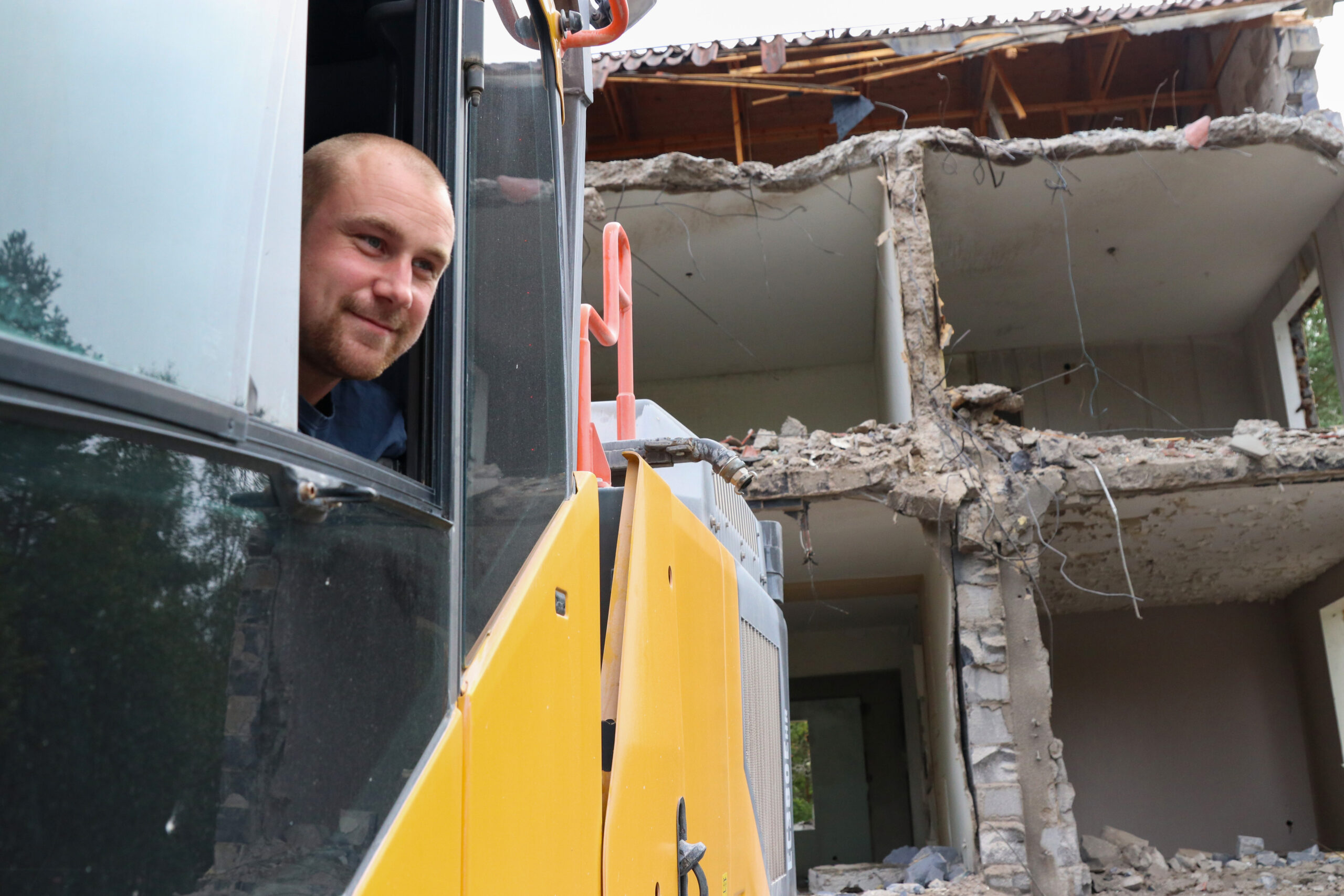 Man som tittar ut från fönstret i en grävmaskin. Bara delar av grävmaskinen syns. I bakgrunden ser man skalet av ett hus.