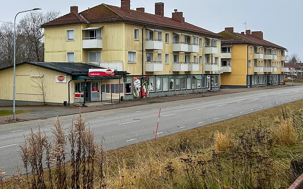 Bilden visar en väg och gula flerfamiljshus. I ett husen syns en nedlagd affär. I anslutning till det första huset syns en nedlagd liten affär.
