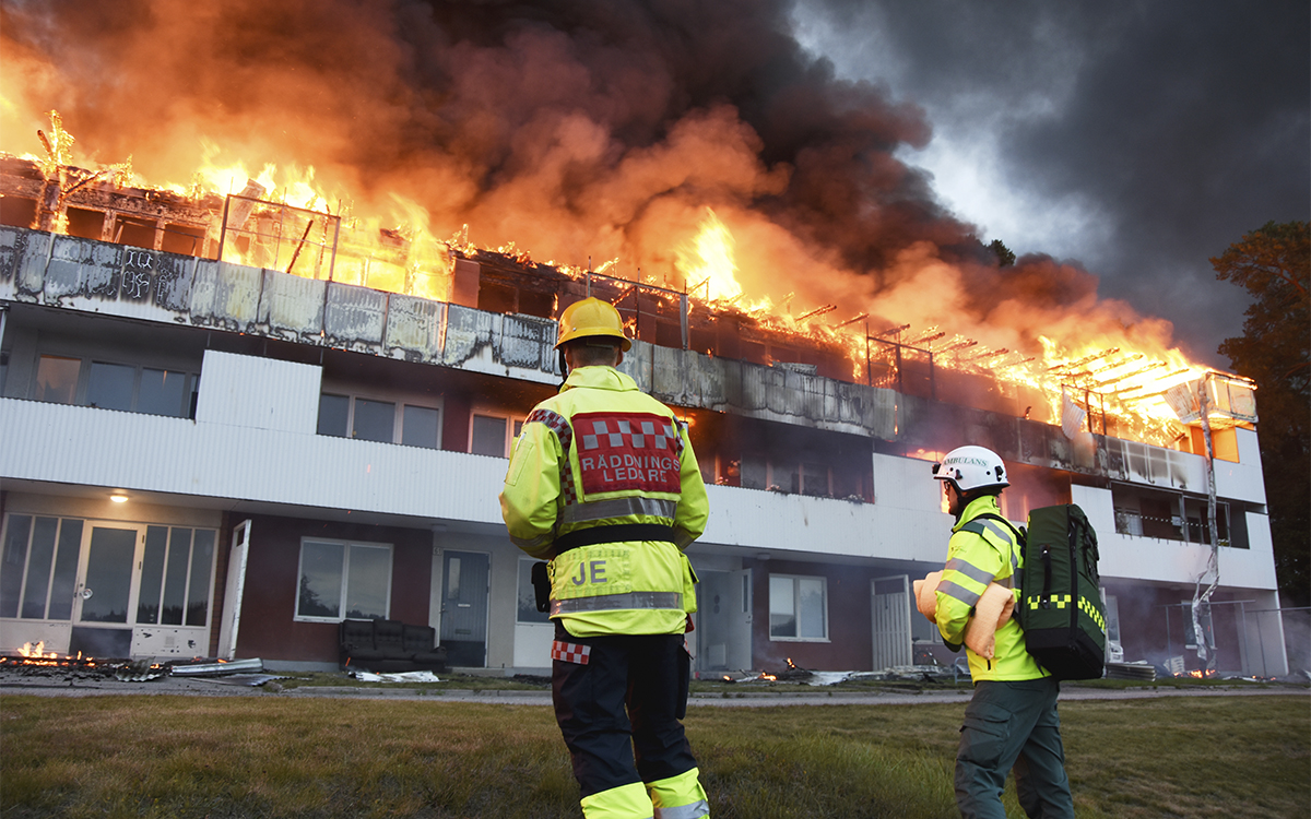 En huslänga i tre våningar brinner med stora lågor längst hela taket och svart rök. Två personer i gula arbetskläder och hjälmar står och tuttar mot branden. Den ena har en röd märkning på ryggen av jackan där det står "räddningsledare". Den andra har en ryggsäck på ryggen och hålller en ihoprullad orange filt under armen. Branden i Söderala 2020 spred sig via vinden som saknade uppdelning i brandceller. Ingen vet hur vanligt det är med flerfamiljshus där vinden saknar brandceller.