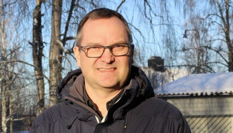 En glasögonprydd man i övre medelåldern tittar in i kameran. Bilden är tagen utomhus.