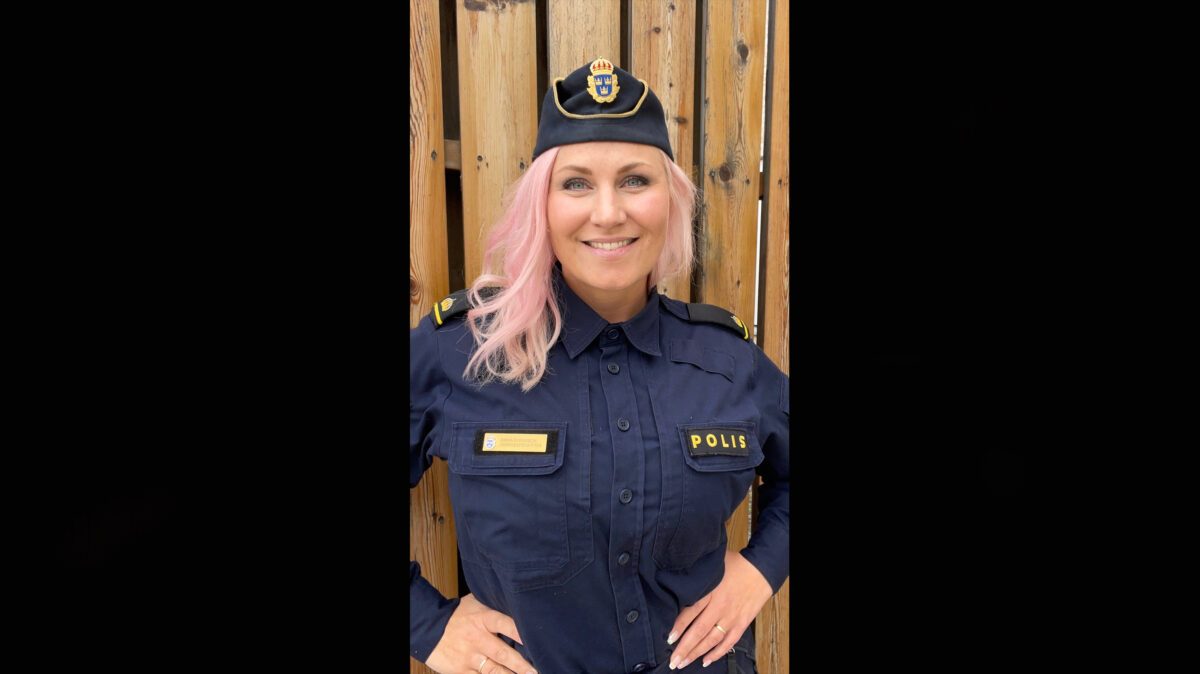 En ung polistjej i uniform. Bilden är tagen utomhus mot ett plank.