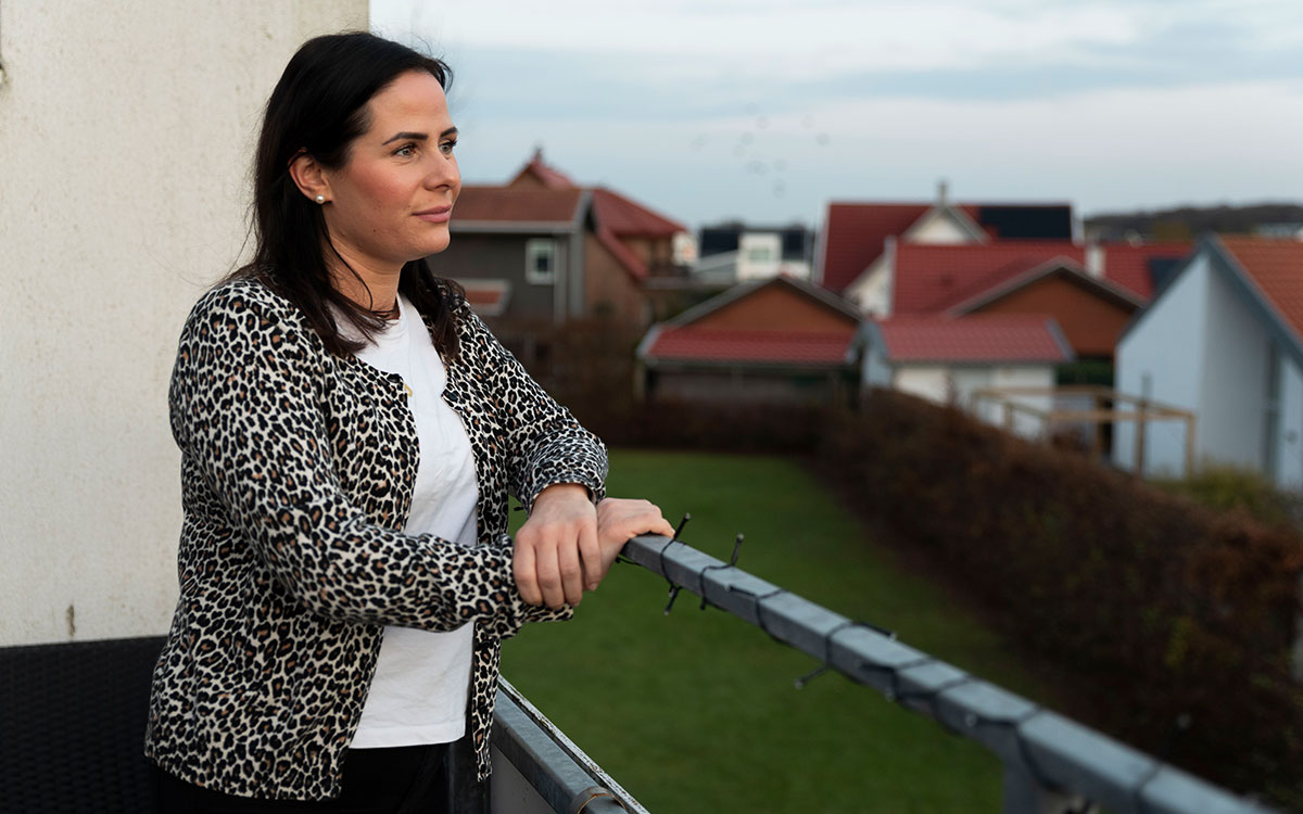 Ett porträtt av en ung kvinna med brunt hår, leopardmöstrad kofta och vit tröja som står på en balkong med hus i bakgrunden.