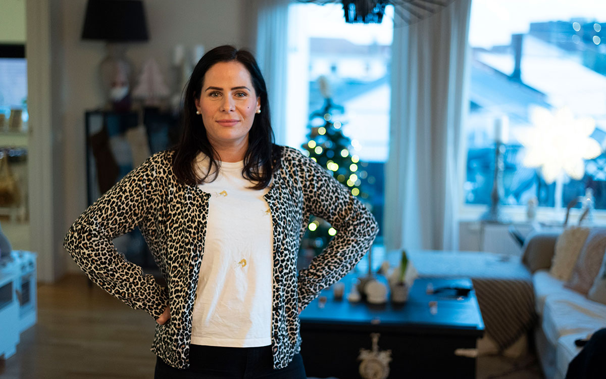 En ung kvinna med brunt hår, leopardmöstrad kofta och vit tröja som står med armarna i sidan i ett vardagsrum.