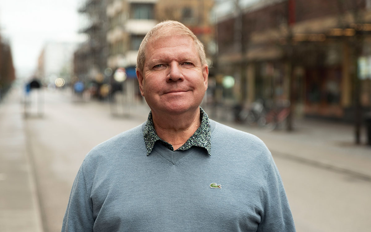 En man med ljust hår och en ljusblå tröja som står utomhus på en gata i en stad.