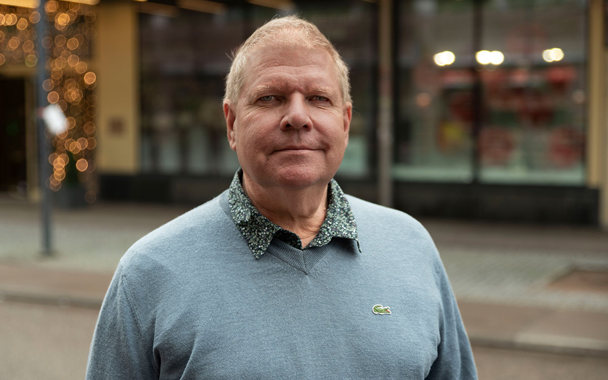 En man med ljust hår och en ljusblå tröja som står utomhus på en gata i en stad.