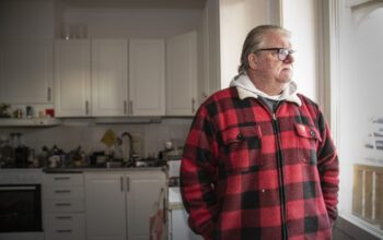 En gråhårig man i pensionsåldern står och tittar ut genom ett fönster i köket. Han bär glasögon, mustasch och en röd- och svartrutig flanellskjorta.