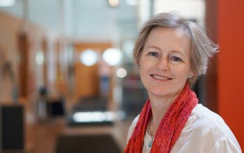 Kvinna som tittar in i kameran och ler. Huvudet och lite av överkroppen syns. Bakgrunden är suddig.