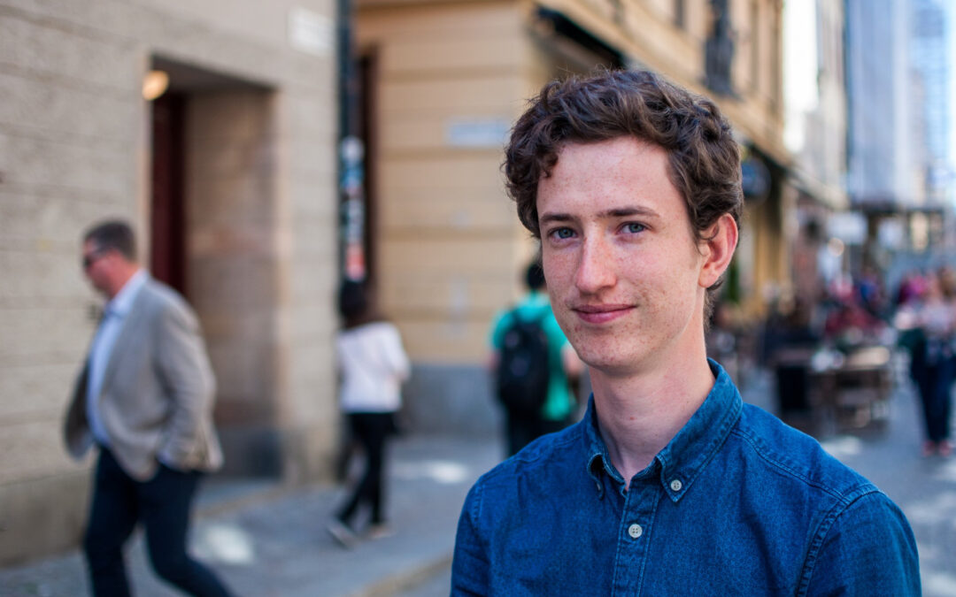 En ung man står på en gata till höger i bilden. Han syns i halvkroppsformat. Han har tjockt brunt kortklippt hår kammat åt sidan. I bakgrunden en gata där det rör sig människor.