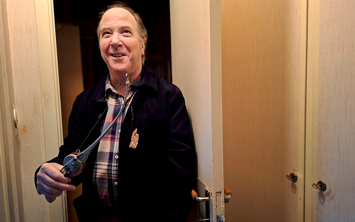 En man som tittar snett upp mot taket och ler. I ena handen håller han ett pimpelspö vars krok är agnat med en bit bröd. Han står i en hall.