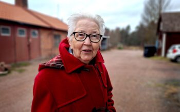 Kvinna som står på en gård där en låg tegelbyggnad skymtar i bakgrunden.