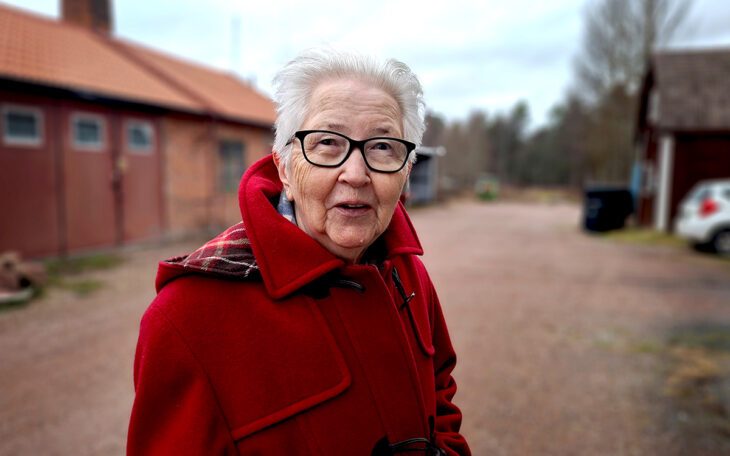 Kvinna som står på en gård där en låg tegelbyggnad skymtar i bakgrunden.