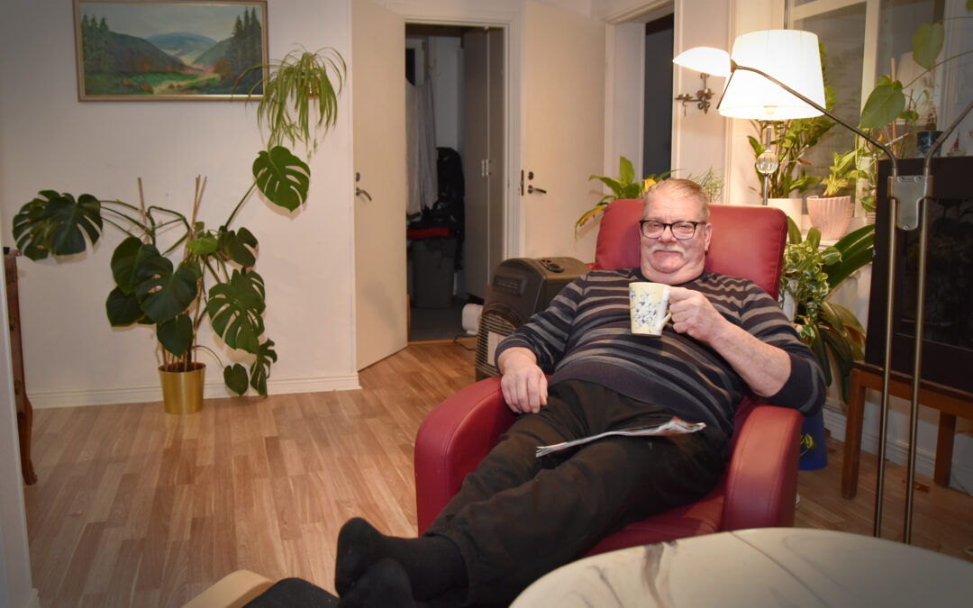En man i pensionsåldern ligger i en fåtölj med fötterna vilandes på en pall och en kaffekopp i handen. Han sitter i ett vardagsrum med stora gröna växter och liten läslampa.