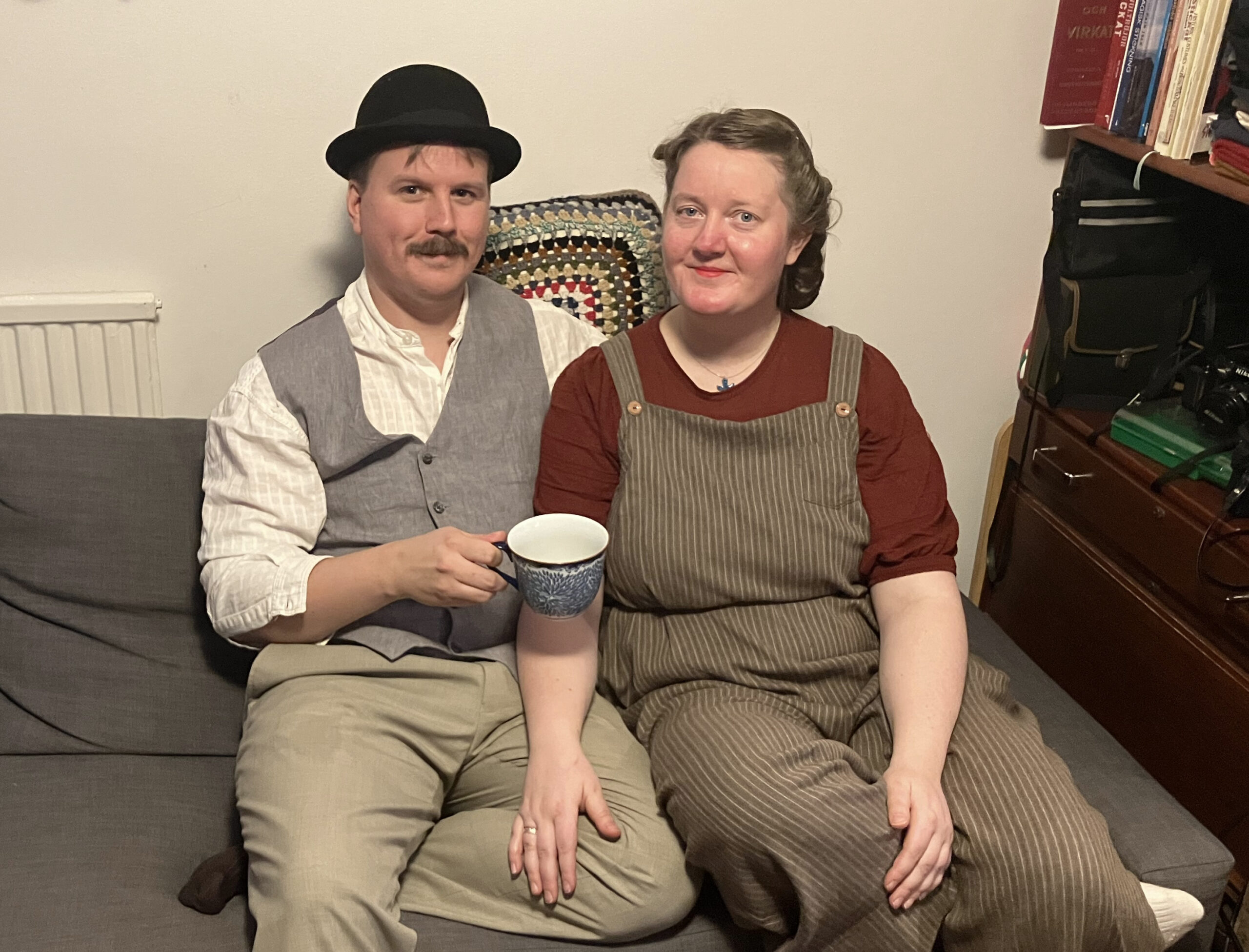 Ett par runt 30 i en soffa. Till vänster en man i svart plommonstop, vit skjorta, grå väst och ljusa byxor. Han har mustasch. I handen håller han en kaffekopp. Kvinnan till höger har håruppsättning i 40-talsstil, vinröd t-shirt och beige hängselbyxor.