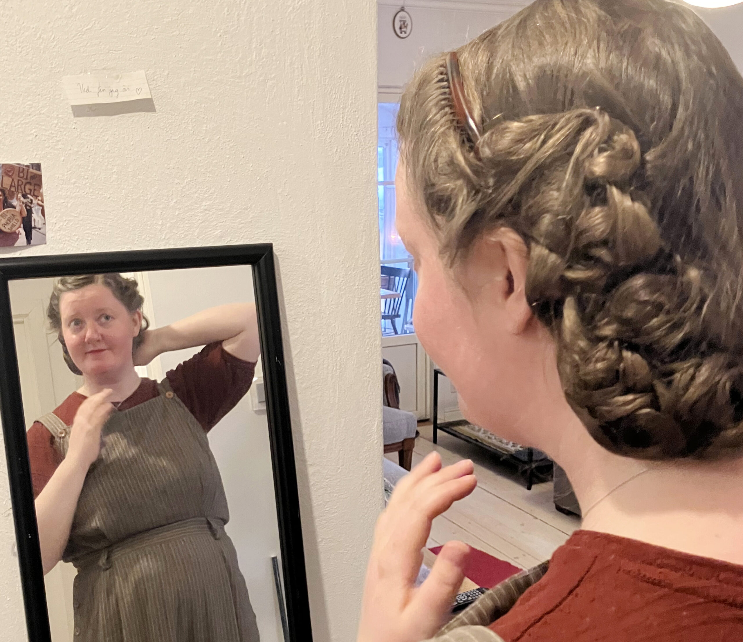 Kvinna speglar sig . Hon har vinröd topp, beige hängselbyxor i linne och en avancerad håruppsättning med kammar, hårnät och lockar.