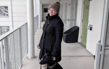 En kvinna i 30-årsåldern står på en loftgång. Hon har svart jacka, beige mössa och glasögon. Hon tittar in i kameran. Vid sin sida har hon en svart mellanstor hund i koppel.