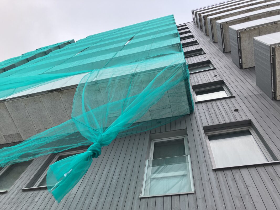 Vy underifrån av balkongen på en 11-våningsbyggnad med grå fasad. Balkongen är täckt med grönt nät.