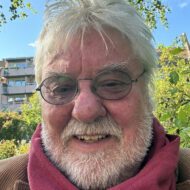 Porträttbild på en äldre man med rufsigt vitt hår, mustasch och kort skägg. Han har tonade glasögon, röd halsduk och beige kappslag. Han ser lätt mot kameran.