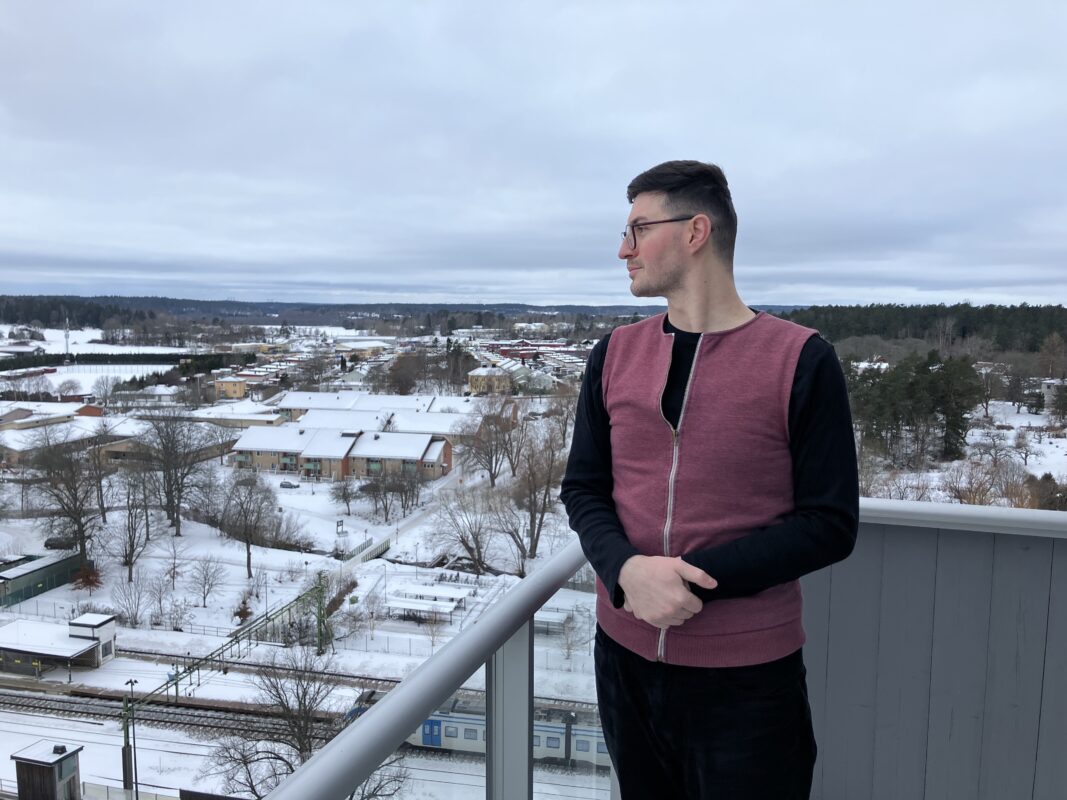 En man i 30-årsåldern står i profil och tittar ut från sin balkong på 11:e våningen. Han har en kort mörk frisyr och glasögon. Han bär en knallröd väst, svart långärmad skjorta och svarta byxor. Nedan kan du se snötäckta tak, kala träd och en skog i bakgrunden. 