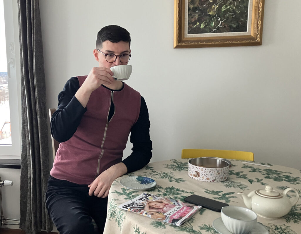 En man med kort svart hår och svarta glasögon dricker te vid köksbordet. Han bär en knallröd väst, svart långärmad skjorta och svarta byxor. En bild hänger på väggen bakom honom. På bordet står ytterligare en tekopp, en tekanna, en kakburk och en tidning som heter Hem & Haila. Blommig duk.